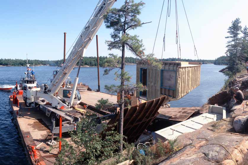 The prefabricated sauna slung from a crane on a barge, swinging over the water toward the granite shore during installation.