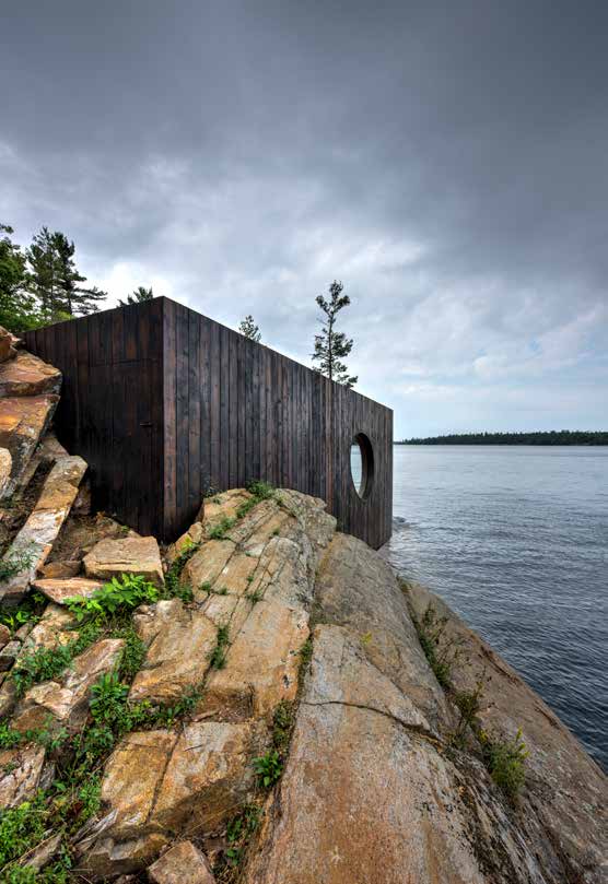 The blackened cedar exterior standing on bare granite under a heavy grey sky, the oval window cut through the wall above the water.