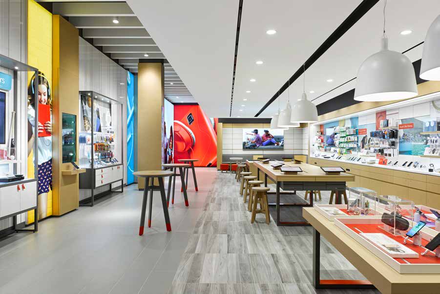 A bright retail store with pale timber display tables running down the centre aisle, backlit product walls to either side and a row of white dome pendants overhead.