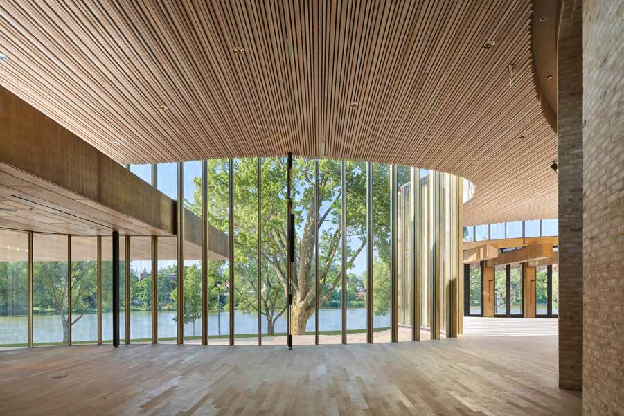 A theatre lobby whose slatted wood ceiling fans out above a curved glass wall of slender mullions, looking across a lawn to trees and the river beyond.